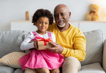 Cheerful small african american girl sits on knees of aged man and holds gift from her grandfather