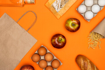 Paper eco bag, bottle of olive oil, eggs, bread, beans, red apple, spiral paste on orange background. Healthy food, delivery, donation concept. Food stock for quarantine. Top view, flatly.