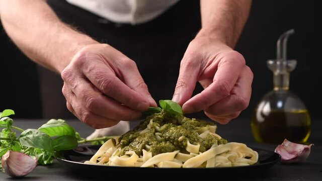 the chef decorates with basil leaf the pasta dish with genovese pesto sauce on black table with sauce ingredients