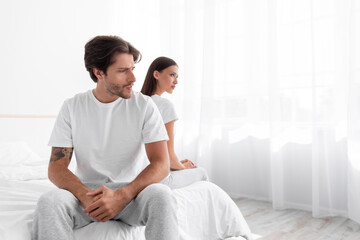 Unhappy upset offended caucasian millennial husband ignores wife sitting in bed in white bedroom interior