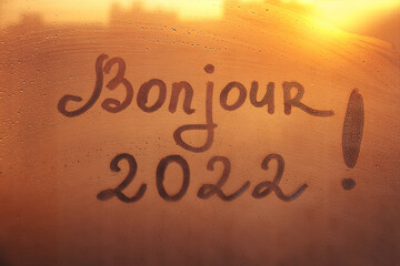 lettering Bonjour in French is hello in english and numbers 2022 paint with finger with streaks of water on splashed by rain foggy glass on orange sunset window © ksenija1803z