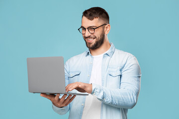 Smiling attractive adult european male in glasses typing on computer, works remotely, isolated on blue background