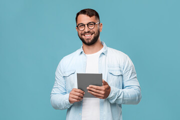 Cheerful mature caucasian guy in glasses with tablet look at camera, isolated on blue background,...