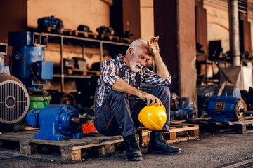 An old, tired bearded factory worker in overalls is sitting on the pallet and taking a break from hard work. He is wiping sweat from his forehead. - obrazy, fototapety, plakaty