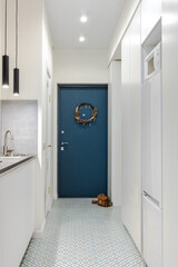 A minimalistic interior of a hallway in a studio apartment in white and gray shades with a blue entrance door. Built-in storage and compact kitchen layout