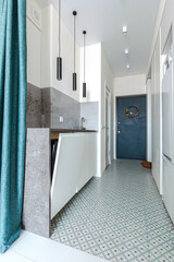 A minimalistic interior of a hallway in a studio apartment in white and gray shades with a blue entrance door. Built-in storage and compact kitchen layout