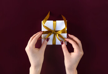 Nice female hands holding gift box wrapped in white paper with golden ribbon on festive dark background. Xmas and New Year postcard design. Black Friday sales, Birthday celebration party concept