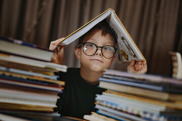 cute caucasian boy wearing glasses covering head with book. Cheerful boy peeking from behind piles...