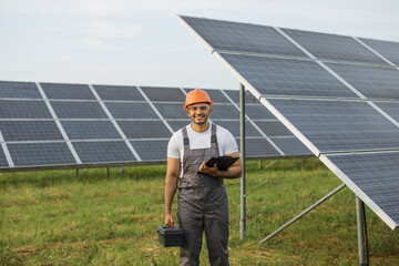 Indian man in uniform, yellow hard hat and safety glasses using clipboard and instruments for work on solar farm. Competent energy engineer controlling work of photovoltaic cells.