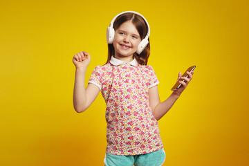 Cheerful young girl with white bluetooth headphones, listening music from smartphone and dancing,...
