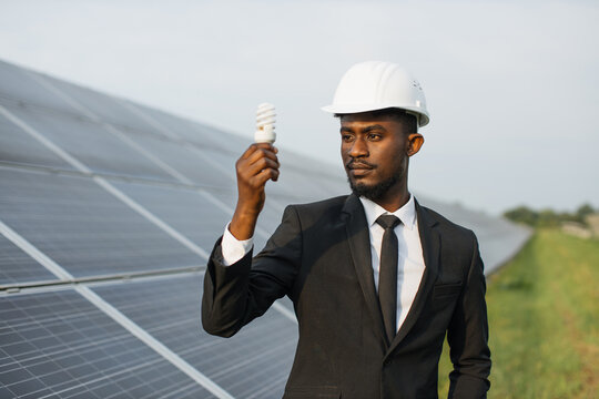 African american man with light bulb in hand standing on field with rows of solar panels. Engineer wearing black suit and white helmet. Concept of alternative energy and environment.