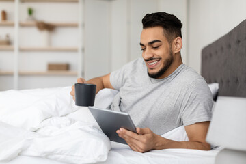 Happy Young Arab Man Relaxing In Bed With Digital Tablet And Coffee