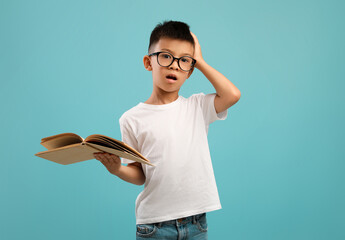 Tired Of Study. Stresses Little Asian Boy Holding Book And Touching Head - obrazy, fototapety, plakaty