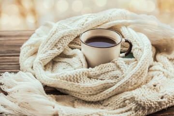 Morning coffee. A mug of hot warming drink in a warm scarf on a wooden background. Cozy autumn-winter concept. Blurred bokeh in the background.