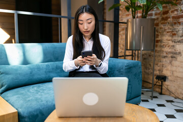 Naklejka premium Asian woman doing online shopping using the phone while sitting at the table in front of the laptop