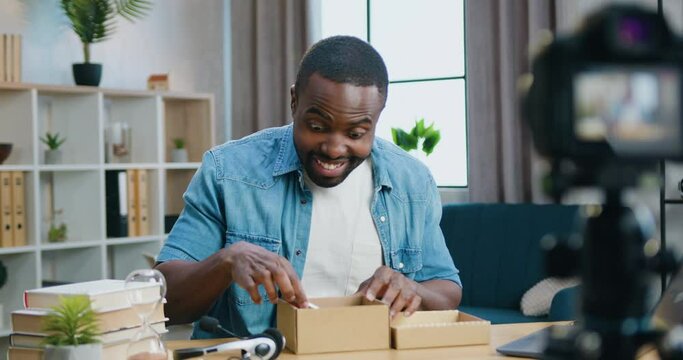 Good-looking happy smiling bearded african american blogger doing big eyes from surprise when opening box and taking out new phone during streaming on camera from home for online spectators