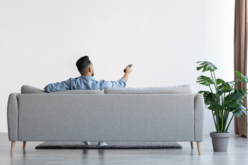 Young Man Sitting On Couch And Opening Air Conditioner With Remote Controller