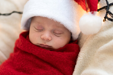 Top View Portrait First Days Of Life Newborn Cute Funny Sleeping Child Baby In Santa Hat Wrapped In Red Diaper At White Garland Background. Merry Christmas, Happy New Year, Infant, Childhood, Winter