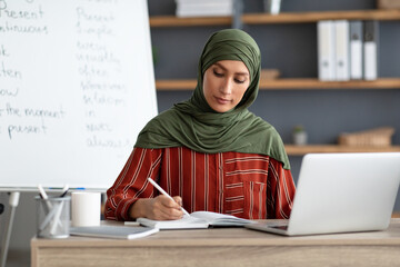 Muslim teacher in headscarf sitting at desk, writing, using pc