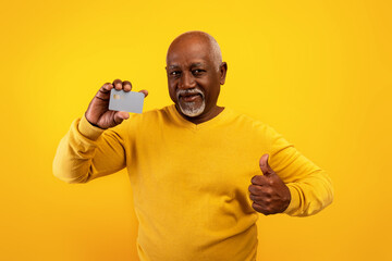 Senior African American man holding credit card and showing thumb up on orange studio background