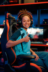 Happy African female cybersport gamer wearing headset showing approval sign while sitting on gaming...