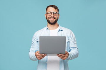 Cheerful adult european male in glasses with laptop looks at camera, isolated on blue background