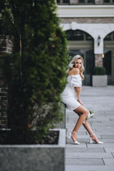 Fototapeta premium A beautiful, young blonde bride in a white dress with a bouquet of peonies sits near the green thuja, cypress on the background of buildings on the street in the city. Wedding photography.