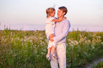 Little caucasian daughter in father's arms in the field on sunset summer time