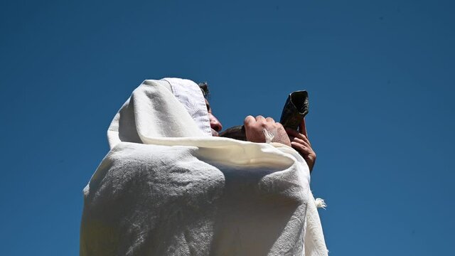 Adult Jewish man blow Shofar (ram's horn) outdoors under clear blue sky, on the Jewish High Holidays in Rosh Hashanah and Yom Kippur.
