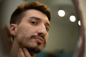 A guy with a patchy beard looks in the mirror