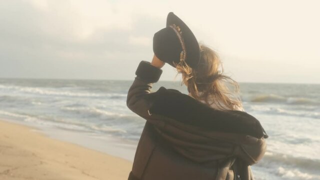 Portrait of romantic brunette woman in hat and leather jacket walking on the beach in autumn at sunset or sunrise 