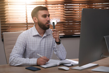 Handsome young man using disposable electronic cigarette at table in office