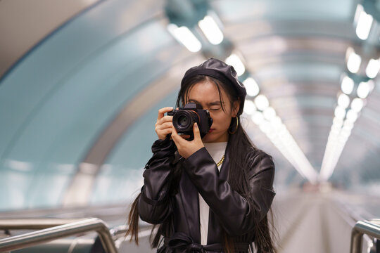 Young stylish asian girl make photos on camera in urban city transport riding escalator. Modern japanese female photographing while travelling. Creative chinese artist making pictures at underground