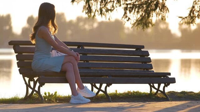 Lonely woman sitting on lake side bench enjoying warm summer evening. Solitude and relaxation concept