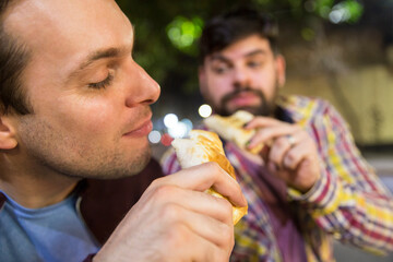 Attractive latin gay couple enjoying a leisure time while eating empanadas outdoors at night.