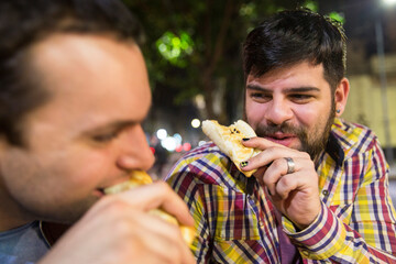 Attractive latin gay couple enjoying a leisure time while eating empanadas outdoors at night.
