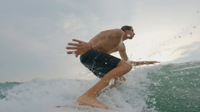 A beautiful Caucasian surfer actively rakes the wave starts and rides it. A young guy is surfing in the ocean and doing performance maneuvers on a white short board. The camera is fixed on the board.