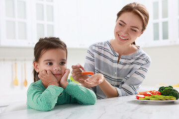 Mother feeding her daughter in kitchen. Little girl refusing to eat vegetables