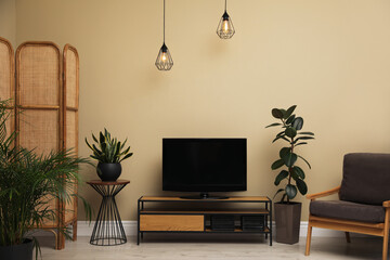 Modern TV on cabinet, armchair and green plants near beige wall in room. Interior design