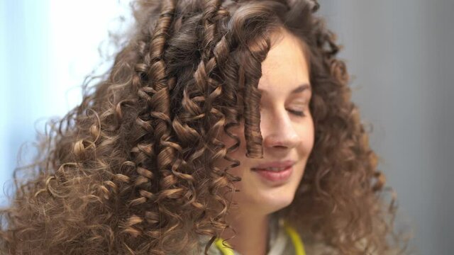 Close-up of a young woman with curly hair who is doing her hair, she laughs looking at the camera, turning her head in different directions. High quality 4k footage