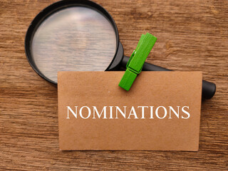 Magnifying glass and brown card with text NOMINATIONS on wooden background.