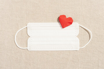 Flat lay of Red heart shape origami paper on white medical face mask on beige linen cloth in soft natural light. Health care, new normal, safety and protection during Covid 19 background concept.