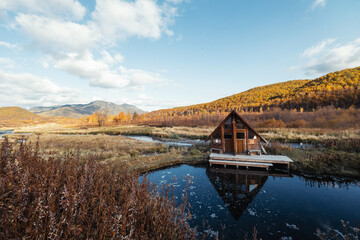 Wooden house near hot spring. First frost, steam over water, beautiful colourful autumn landscape