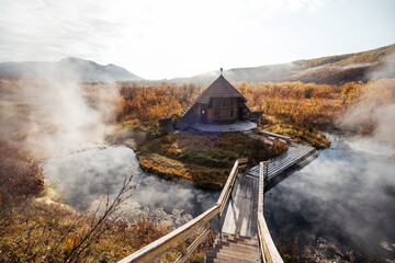 Wooden house near hot spring. First frost, steam over water, beautiful colourful autumn landscape