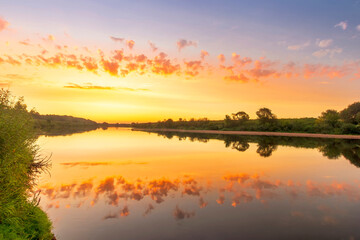 Naklejka na ściany i meble Scenic view at beautiful summer river sunset with reflection on water with green bushes, grass, calm water ,deep cloudy sky and glow on a background, spring , evening landscape