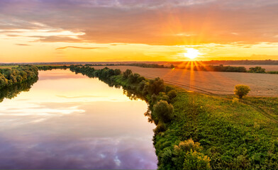 Fototapeta na wymiar Scenic view at beautiful summer river sunset with reflection on water with green bushes, grass, golden sun rays, calm water ,deep cloudy sky and glow on a background, spring , evening landscape