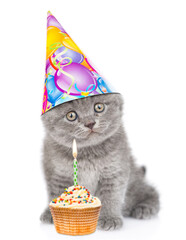 Kitten wearing birthday cap sits cup cake and looks at camera. isolated on white background