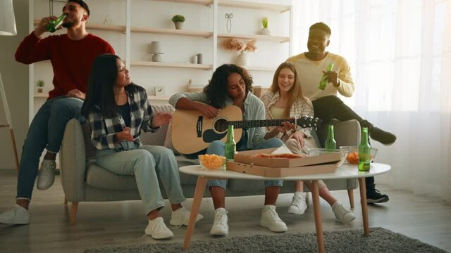 Cute young diverse friends playing guitar and singing songs together, having party at home, sitting on couch in living room