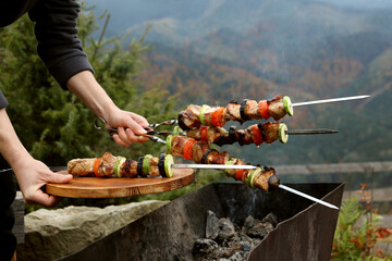 Woman holding metal skewers and board with delicious meat against mountain landscape, closeup