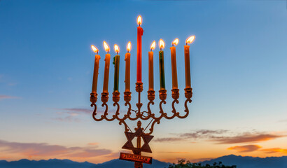 Burning festive candles are traditional symbols of Hanukkah Hebrew Holiday of Light, selective focus on menorah with burning  wax candles, blurred mountains and dramatic dawn sky background 